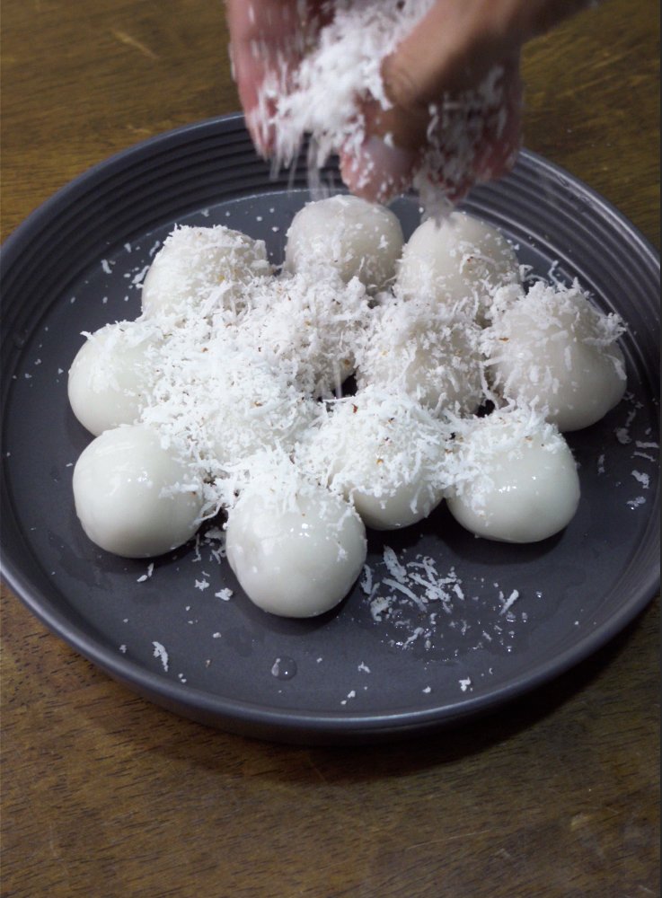 Mont lone yay paw, white sticky rice balls on a dark plate being sprinkled with grated coconut